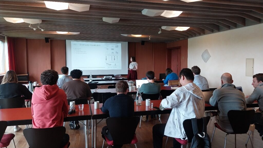 PhD presentation at the doctoral retreat. A group of people can been from behind sitting at three rows of desks with some space between them. A young woman is standing in front of a projection screen at the front of the room. On the screen, there are a few graphics and the text "Uncertainty Visualization" 
