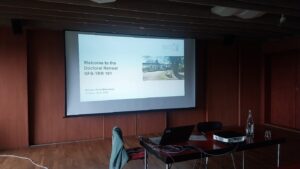 Welcome presentation for the doctoral retreat. On a projection screen, the opening slide of a presentation reads "Welcome to the Doctoral Retreat SFB-TRR 161". In front, there is a desk with an open laptop and a jacket thrown over an empty chair. Both the floor and the walls of the room are paneled with dark wood.