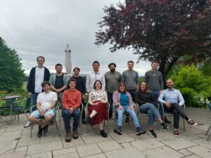 Group of Doctoral Students who participated in the doctoral retreat. 6 people are sitting on chairs in the front row, 7 people are standing behind them. There are three women sitting in the front row, the rest of them are men. All are dressed casually and are smiling for the camera. In the background, there is a tree with purple leaves, a folded sun-umbrella, and a grey sky.