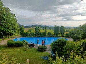 View from the Humboldt haus at the Doctoral Retreat. A group of five people can be seen from distance, some of them standing, some sitting on a bench. Behind them, a swimming pool is located on a terrace. The area is surrounded by lush greenery and in the background some hills and forests are visible. The sky is overcast with grey clouds.