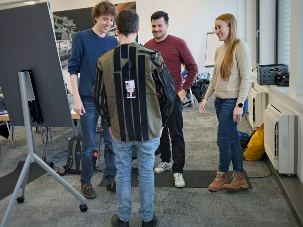 Three participants of the research hackathon test their prototype. A mobile phone is taped to the back of a man. Two other men and one woman are standing in front of the man.