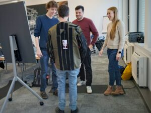 Three participants of the research hackathon test their prototype. A mobile phone is taped to the back of a man. Two other men and one woman are standing in front of the man.
