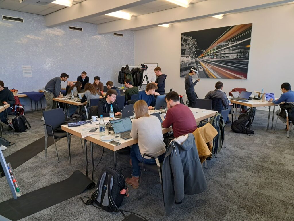 Participants of the research hackathon can be seen sitting in groups of 4 to 8 people at tables, most of them working on their laptops.